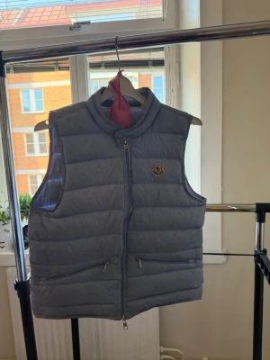 Blå moncler gui väst - Snygg blå gui väst  från Moncler med hög krage och två dragkedjefickor framtill. Västen har quiltad design och Moncler-logga på bröstet. Västen är lite skitig, men efter en tvätt är den som ny