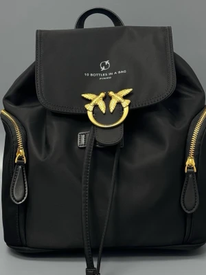 Från Pinko Svart ryggsäck  med gulddetaljer - Stilren svart ryggsäck från Pinko i nylon med exklusiva guldfärgade metalldetaljer, inklusive den ikoniska fågelspännet framtill. Väskan har två sidofickor med dragkedja och justerbara axelband. Texten '10 Bottles In A Bag' tryckt på locket. Mått 26/27 cm nypris är ca 5000kr