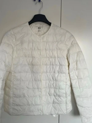 Vit dunjacka från Uniqlo - Stilren vit dunjacka från Uniqlo med quiltat mönster och rund halsringning. Lätt och smidig modell med diskret knäppning framtill. Perfekt att slänga över en hoodie eller tröja för extra värme. Klassisk och enkel design som funkar till det mesta.