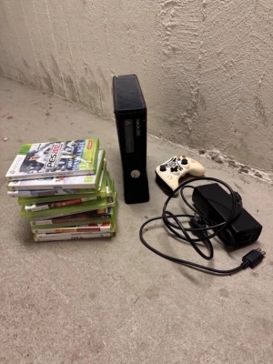 Xbox 360 Slim med spel och tillbehör - Säljer en svart Xbox 360 Slim från Microsoft med en vit handkontroll, strömadapter och flera spel. Konsolen har en stilren, rektangulär form och är tillverkad i plast. Perfekt för gaming med kompisar eller solo.