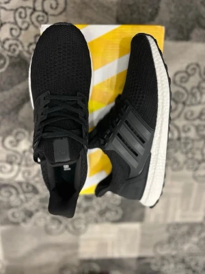 Adidas Ultra Boost svarta sneakers - Svarta Adidas Ultra Boost sneakers med stickad ovandel och klassiska tre ränder i plast på sidorna. Skorna har en vit Boost-sula för extra komfort och svart yttersula. Perfekta för dig som gillar stilrena och sportiga sneakers.