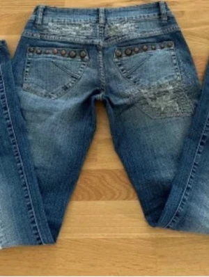 Blå bootcut jeans med nitar - Säljer ett par blå bootcut jeans med slitningar och dekorativa nitar på bakfickorna. Jeansen har klassisk femficksdesign och är tillverkade i denim.  Innerbenslängd: 79 cm Midjemått tvärs över 36 cm Gylfmått: 9 cm 