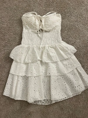 Vit volangklänning med spetsdetaljer - Supersöt vit kortklänning med volanger och broderade spetsdetaljer. Klänningen har smockad midja, knytning framtill och tunna axelband. Perfekt för sommarens alla tillfällen och ger en romantisk vibe.