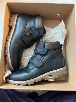 Marinblå kängor med kardborre från Ecco - Snygga marinblå kängor från Ecco i läder med två breda kardborreband och grov beige sula. Skorna har vadderad ankel och en stilren design som passar till många outfits. Perfekta för höst och vinter när du vill ha både stil och funktion.