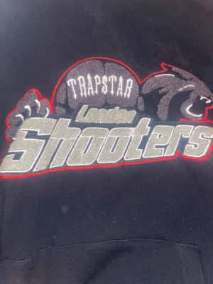 Trapstar London Shooters hoodie svart - Snygg svart hoodie från Trapstar med stort broderat motiv på bröstet där det står 'Trapstar London Shooters' i grått och vitt med röda detaljer. Hoodien har en klassisk streetwear-look och är perfekt för dig som gillar bold prints och unik design.