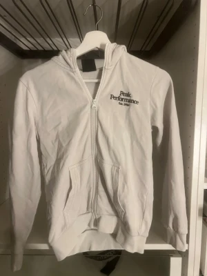 Vit Peak Performance hoodie - Snygg vit hoodie från Peak Performance med dragkedja framtill och broderad logga på bröstet. Klassisk modell med huva och fickor på magen. Perfekt för en avslappnad och sportig stil.