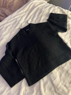 Svart stickad tröja från Gina Tricot - Säljer en svart stickad tröja från Gina Tricot med rund halsringning och breda ribbade muddar vid ärmslut och nederkant. Tröjan har en klassisk, rak passform använt en gång och köptes för 300 och säljer på grund av att den inte används 