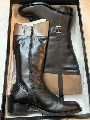 Svarta och beige stövlar Catwalk - Snygga knähöga stövlar från Catwalk Stockholm i svart och beige läder. Stövlarna har dragkedja på sidan, dekorativt spänne och låg klack. Perfekta för dig som vill ha en stilren och trendig look.