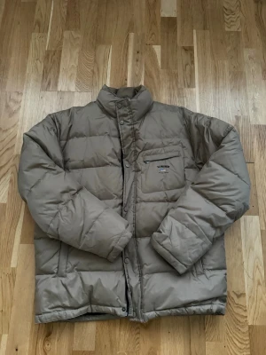 Beige pufferjacka - Varm och rejäl pufferjacka från U.S. Polo Assn. i en mycket snygg och trendig beige/brun nyans. Jackan har hög krage, dragkedja samt knappar och sidofickor. Perfekt för höst och vinter.