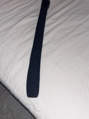 Mörkblått flätat bälte - Snyggt mörkblått flätat bälte med silverfärgat spänne. Bältet har en stilren design och passar perfekt till jeans eller chinos. Materialet är elastiskt syntet som ger en bekväm känsla och bra passform.