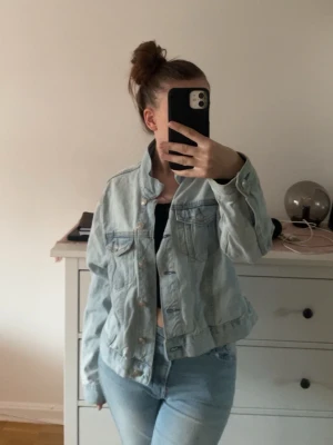 Ljusblå jeansjacka med klassisk passform - Säljer en ljusblå jeansjacka med klassisk passform och snygga detaljer som bröstfickor med knappar och silverfärgade knappar framtill. Jackan är tillverkad i denim och har en cool, avslappnad look som passar perfekt till jeans eller kjol.