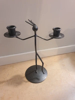 Ljusstake i metall stringfigur - Ljusstake i metall formad som en figur. Ljusstaken har två hållare för ljus, en på varje sida om figuren. Figuren står på en rund bas. Den verkar vara tillverkad i Danmark är inte säker då jag inte vet när och var den införskaffats.