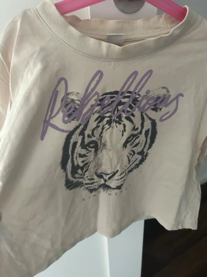Beige crop t-shirt med tigertryck - Säljer en beige crop t-shirt från Lindex med ett stort tigertryck och texten 'Rebellious' i lila framtill. T-shirten har rund halsringning och är tillverkad i mjuk bomull. Perfekt för dig som gillar djurmotiv och en cool stil.
