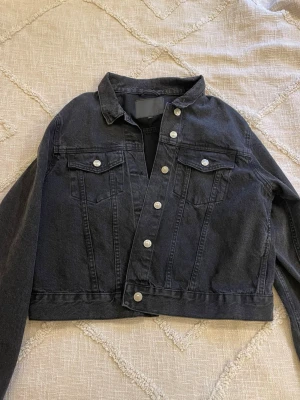 Svart croppad jeansjacka - Snygg svart croppad jeansjacka med klassisk krage och silverfärgade knappar. Två fickor med lock och knapp framtill. Rak passform och långa ärmar. Säljer pågrund av att jackan inte kommit till användning.