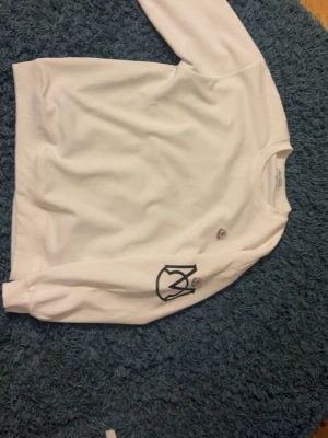 Vit sweatshirt - Säljer en vit sweatshirt  med rund halsringning och svart tryckt logga på ena ärmen. ribbade muddar vid ärmslut och nederkant. Perfekt för en clean och stilren look. Storlek L men sitter som m