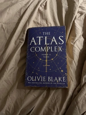 The Atlas Complex av Olivie Blake - En engelskspråkig skönlitterär bok med blått omslag och guldiga detaljer. Titeln är 'The Atlas Complex' och författaren är Olivie Blake. Omslaget har stjärnmönster och en gyllene symbol, vilket ger boken en mystisk och elegant känsla.