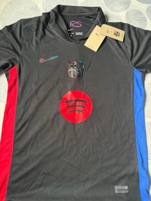 Svart FC Barcelona Nike matchtröja - Snygg svart FC Barcelona matchtröja från Nike med Dri-FIT material. Tröjan har klubbmärke, Nike-logga och Spotify-logga på bröstet samt röda och blå detaljer på sidorna. Perfekt för dig som älskar fotboll och vill ha en unik supportertröja.