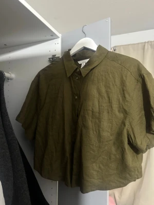 Olivgrön croppad linneskjorta - Säljer en olivgrön croppad skjorta från H&M i linne. Skjortan har korta ärmar, klassisk krage och knappar framtill. Perfekt för dig som gillar en avslappnad och trendig stil. Passar som s 