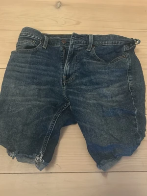 Blå jeansshorts med rå kant - Säljer ett par klassiska blå jeansshorts med rå, fransig kant nertill. Shortsen har fem fickor, bälteshällor och knappgylf. Perfekta för sommaren och har en avslappnad passform. Inget synligt märke på shortsen. Storlek: W31.