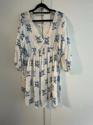 Vit blommig omlottklänning - Söt kortklänning i vit bomull med blått blommönster. Klänningen har omlottmodell med v-ringning, trekvartsärmar och knytband i ryggen. Perfekt för sommaren!