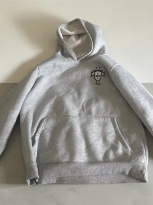 Grå Portugal hoodie - Snygg grå hoodie med ett svartvitt tryck på bröstet. Klassisk känguruficka framtill och ribbade muddar vid ärmslut och nederkant. Mjuk insida och avslappnad passform, perfekt för en chill vardagslook. Obs passar även M.