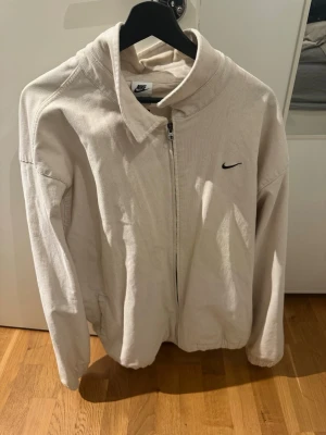 Beige zipjacka från Nike - Snygg beige zipjacka från Nike med diskret svart logga på bröstet. Jackan har krage, dragkedja framtill och resår i midjan. Tillverkad i ett mjukt, ribbat tyg som ger en avslappnad och sportig look. Perfekt för lager-på-lager-stil.