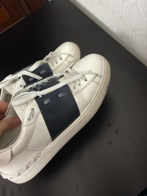 Valentino - Snygga vita sneakers med bred svart rem över sidan och coola nitar runt hälen. Skorna har vita snören och en chunky sula. Perfekta för dig som gillar stilrena men ändå unika detaljer. Passar till det mesta och ger en clean look.