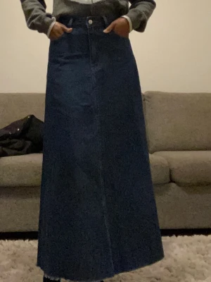 Mörkblå lång jeanskjol - Säljer en mörkblå lång jeanskjol med hög midja och klassisk femficksdesign. Kjolen har en rak, lätt utsvängd passform och stängs med knapp och dragkedja framtill. Perfekt för dig som gillar en enkel men snygg denimlook.