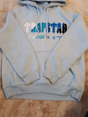 Ljusblå Trapstar hoodie med logga - Säljer en ljusblå hoodie från Trapstar med stor logga i vitt, blått och svart på bröstet. Hoodien har huva med dragsko, magficka och ribbade muddar vid ärmar och nederkant. Mjuk insida i fleece och cool streetwear-vibe.