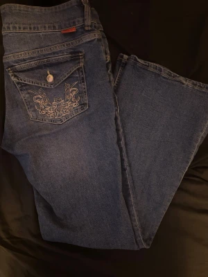 Mörkblå jeans med broderad ficka - Snygga mörkblå jeans från Angelic/H&M med unik broderad design på bakfickan och knappdetalj. Klassisk femficksmodell i med bootcut. Perfekta för dig som vill ha ett par jeans med lite extra attityd.