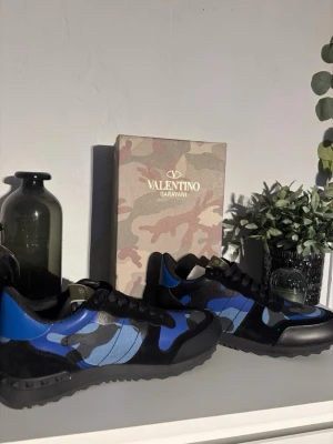 Valentino Garavani blå camo sneakers - Unika sneakers från Valentino Garavani med blått, svart och grått camo-mönster. Skorna har svarta snören, detaljer i mocka och läder samt en robust svart gummisula med nitar på hälen. Perfekt för dig som gillar exklusiv och trendig streetstyle.