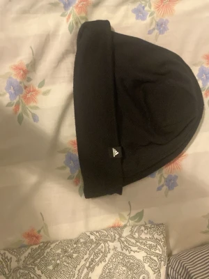 Svart mössa från Adidas - Snygg svart mössa från Adidas med uppvikt kant och diskret logga framtill. Perfekt för kalla dagar och enkel att matcha med olika outfits. Klassisk och stilren design.
