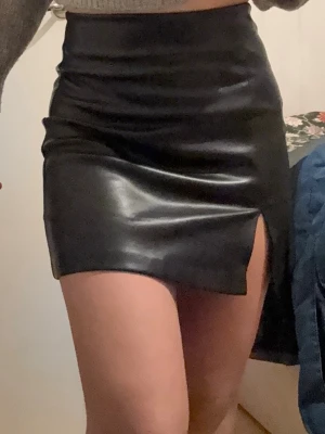 Svart minikjol i skinnimitation - Säljer en svart kortkjol i skinnimitation med slits framtill. Kjolen har en tight passform och en stilren, minimalistisk look som passar perfekt till en trendig outfit.