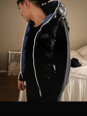 Svart dunväst från Duvetica med huva - Snygg svart dunväst från Duvetica med glansig finish och stor huva. Västen har vit dragkedja och blått innerfoder, samt en diskret logga på bröstet. Perfekt för lager-på-lager och streetwear-stil. Priset är diskuterbart 