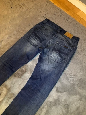 Nudie jeans  - Säljer ett par klassiska blå jeans från Nudie Jeans med raka ben och snygga slitningar på bakfickorna. Jeansen har fem fickor, normal midja och är gjorda i denim av bomull. Perfekta för en avslappnad och trendig look.