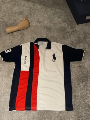 Randig pikétröja från Ralph Lauren - Snygg pikétröja från Ralph Lauren med breda ränder i vitt, marinblått och rött. Tröjan har klassisk krage, korta ärmar och broderad logga på bröstet. Perfekt för dig som gillar sportig och stilren look.