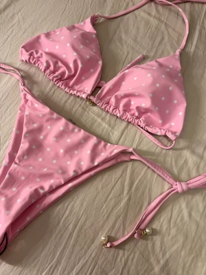 Rosa prickig bikini med knytband - Supersöt rosa bikini med vita prickar och knytband både vid nacken, ryggen och höfterna. Bikinin har små pärldetaljer på snörena och en klassisk trekantsmodell på toppen. Säljer pga köpte fel storlek så den är endast testad💕