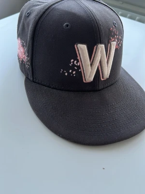 Svart keps med W och körsbärsblom - Säljer en svart keps med stor broderad W framtill och rosa detaljer som liknar körsbärsblommor. På sidan finns ett broderat träd med rosa blommor. Kepsen har rak skärm och klassisk snapback-form. Perfekt för dig som vill sticka ut med snygga detaljer. Storlek i sista bilden👍