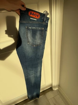 Blå Dsquared2 jeans - Säljer ett par blå Dsquared2 Bros jeans med tydliga slitningar och tvättade detaljer. Jeansen har en klassisk femficksmodell, smal passform och en stor orange Dsquared2 Bros patch bak i midjan. 