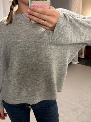 Ljusgrå oversized stickad tröja - Mysig ljusgrå stickad tröja med avslappnad passform och långa ärmar. Tröjan har rund halsringning och ribbade muddar vid ärmslut och nederkant. Litet hål på magen, se bild.