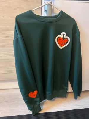 Grön sweatshirt med hjärtlappar - Mörkgrön sweatshirt med rund hals och ribbade muddar. På bröstet sitter en stor applikation i form av ett vitt hjärta med röd detalj, och på ena ärmen finns en mindre röd hjärtlapp. Perfekt för dig som gillar unika detaljer och en chill stil.