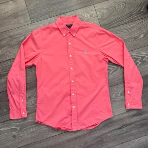 Rosa skjorta från Polo Ralph Lauren - Snygg rosa skjorta från Polo Ralph Lauren med klassisk button-down krage och broderad logga på bröstet. Skjortan är långärmad och har vita knappar framtill samt vid ärmsluten. Perfekt för dig som vill ha en stilren och färgglad look.