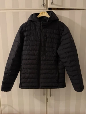 Svart dunjacka Peak Performance - Snygg svart dunjacka från Peak Performance med huva och dragkedja framtill. Jackan har en bröstficka med dragkedja och diskret logga på bröstet. Lättviktig och quiltad design som håller värmen under kalla dagar.