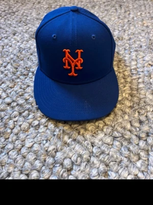 Blå New Era keps med Mets-logga - Säljer en blå keps från New Era med New York Mets broderad logga i orange framtill. Klassisk rak skärm och broderad New Era-logga på sidan. Tillverkad i slitstarkt tyg, perfekt för dig som gillar sportig stil.
