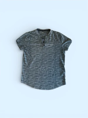  Hollister tröja - Snygg gråmelerad t-shirt från Hollister med korta ärmar och knappdetaljer vid halsen. T-shirten har en liten broderad logga på bröstet och är tillverkad i mjuk bomull. Passformen är slim och tyget har en lätt ribbad känsla.