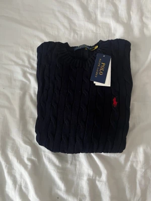 Mörkblå kabelstickad tröja Polo Ralph Lauren - Kabelstickad mörkblå tröja från Polo Ralph Lauren med rund halsringning och klassisk röd broderad logga på bröstet. Tröjan har långa ärmar och ribbade muddar vid ärmslut och nederkant. Perfekt för dig som gillar tidlös och stilren design.