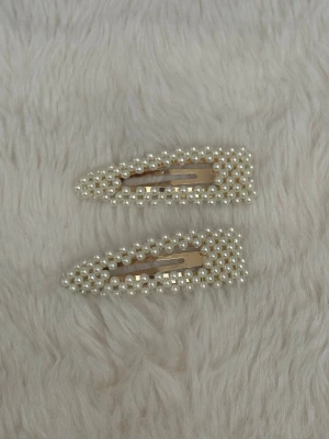 Pärlbeströdda hårspännen - Två stilrena hårspännen dekorerade med små vita pärlor på en guldfärgad metallbas. Spännena har en avsmalnande form och ger en elegant touch till frisyren. Perfekta för att lyfta din look med en trendig accessoar.