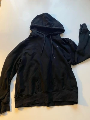 Svart hoodie med tryck 'original' - Svart hoodie med huva och dragsnören. På bröstet finns ett litet vitt tryck med texten 'original'. Hoodien har klassisk passform och ribbade muddar vid ärmslut och nederkant. Perfekt för en avslappnad och stilren look.