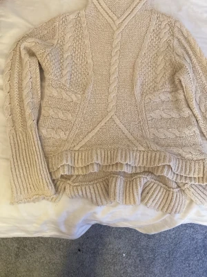 Beige stickad polotröja KappAhl - Säljer en beige stickad polotröja från KappAhl med flätstickade detaljer och ribbade muddar. Tröjan har lång ärm och hög krage, perfekt för kalla dagar. Materialet känns mjukt och mysigt, och designen är både klassisk och trendig.