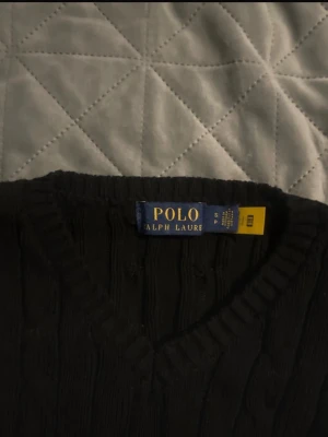 Svart kabelstickad tröja Polo Ralph Lauren - Kabelstickad svart tröja från Polo Ralph Lauren med v-ringning och långa ärmar. Klassisk och tidlös design med ribbade muddar vid ärmslut och nederkant. Perfekt för lager på lager och enkel att matcha med olika outfits.